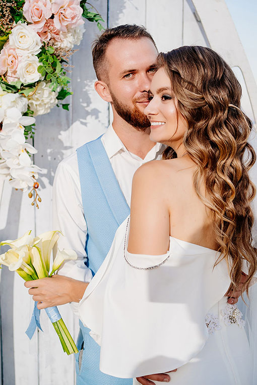 Portrait einer maritimen Hochzeit mit weiß blauen Akzenten. Hübsches Brautpaar posiert vor einem Holzboot mit Blumendekor. Die Braut mit natürlicher Lockenfrisur zeigt ihr rückenfreies trägerloses Hochzeitskleid und hält ihren Brautstrauß aus weißen Callas in der Hand.