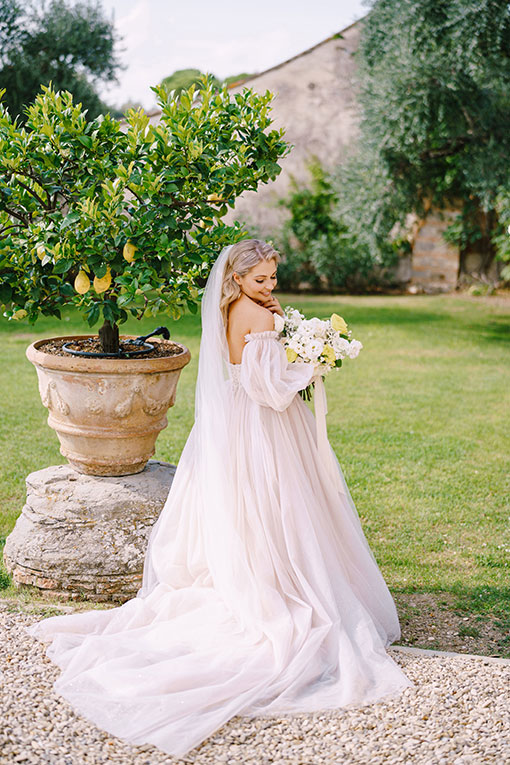 Junge Braut in einem romantischen Hochzeitskleid aus Tüll mit langem Schleier und gelb-weißem Brautstrauß steht im Garten eines alten Weingutes in der Toskana, Italien neben einem Zitronenbaum.