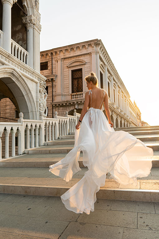 Destination Wedding. Hübsche junge Braut läuft über eine Brücke in Venedig. Ihr rückenfreies weißes Brautkleid fliegt leicht im Wind.