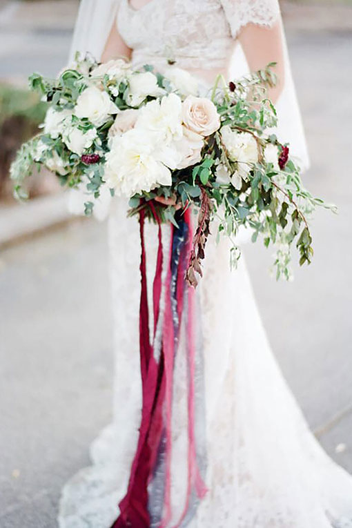 Brautaccessoire. Hochzeitsfloristik für den Brautstrauß. Üppiger Blumenstrauß aus Rosen und Pfingstrosen mit langen, herabhängenden Bändern aus Satin in Bordeaux Rottönen.