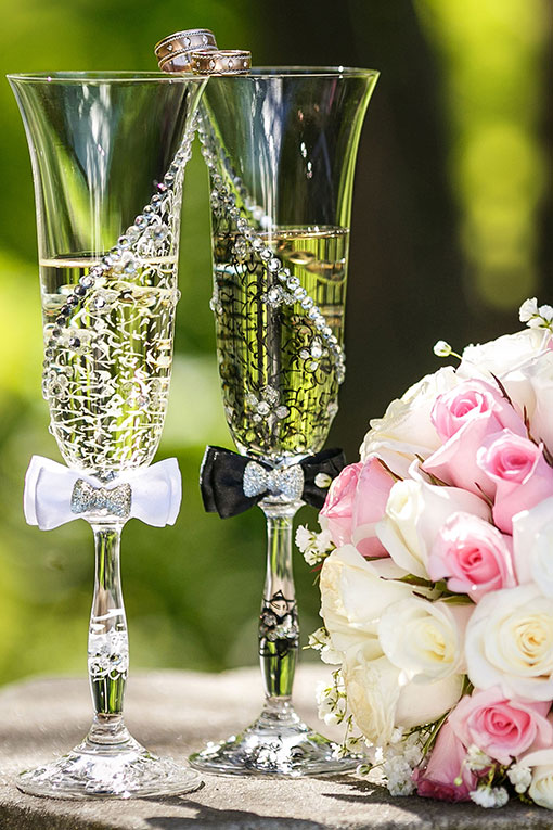 Personalisierte Champagnergläser für das Hochzeitspaar mit Strass, weiße Fliege für die Braut, schwarze Fliege für den Bräutigam. Hochzeitsgläser stehen auf einer Steintreppe mit Trauringen und Brautstrauß.