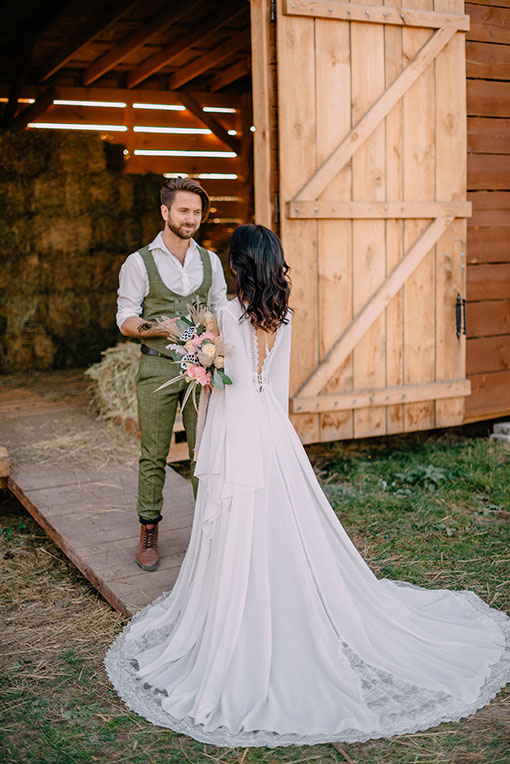 Boho Wedding im Country Style. Trauzeremonie auf einer Ranch. Der Bräutigam steht auf der Rampe zum Heulager und empfängt seine Braut. Er trägt einen lässig stylischen Tweed Herrenanzug in grün mit braunen Stiefeln.