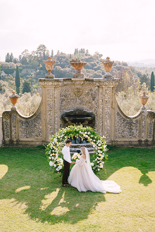 Freie Trauung auf einem alten Weingut in der Toskana, Italien. Hochzeitszeremonie im Garten. Romantisches Brautpaar steht vor einem Traubogen mit weißen und gelben Rosen an einer antiken Mauer mit Amphoren und gibt sich das Ja-Wort.