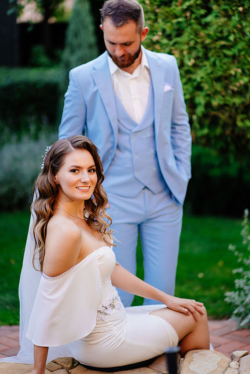 Der Bräutigam trägt einen schicken eleganten Hochzeitsanzug in hellblau mit weißem Einstecktuch. Der Bräutigam steht im Garten hinter der sitzenden Braut in einem schlichten Mermaid Kleid mit Flügelärmeln, Strass und Blütenapplikationen an der Taille.