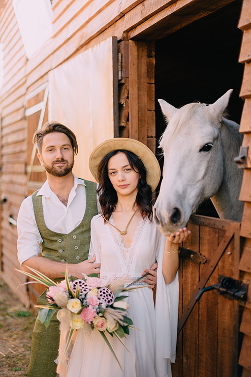 Boho Hochzeit. Fesches Brautpaar im Boho Style posiert vor einer Pferdebox. Ein weißes Pferd blickt heraus.