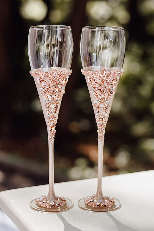 Accessoire für eine Luxushochzeit. Individuell mit Roségold und Strass Steinen verzierte Champagner Gläser für Braut und Bräutigam stehen auf einem weißen Tisch.
