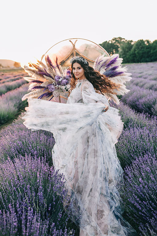 Destination Wedding in Provence. Wunderschöne Braut mit Blumenkranz aus Lavendelblüten und langen Locken tanzt in einem Lavendelfeld vor einem Traubogen, der mit weißen und lila Blüten und Federn dekoriert ist. Hochzeitsvorbereitungen.