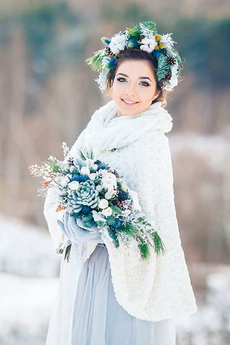 Winter Wedding. Wunderschöne Braut in einem eisblauen Winter Hochzeitskleid und weißen Umhang. Auf dem Kopf Brautschmuck und im Arm Brautstrauß aus winterlichen Blumen und Tannenzapfen.
