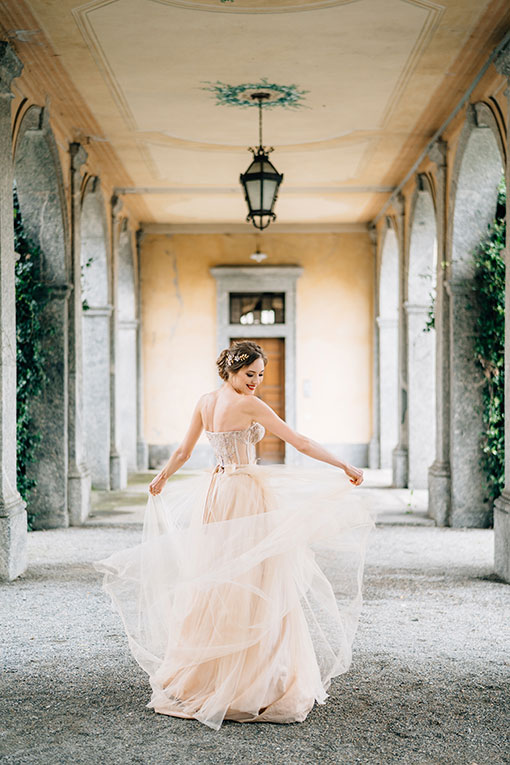 Strahlende junge Braut wirbelt ihr Hochzeitskleid tanzend in einem alten Säulengang im Grünen.
