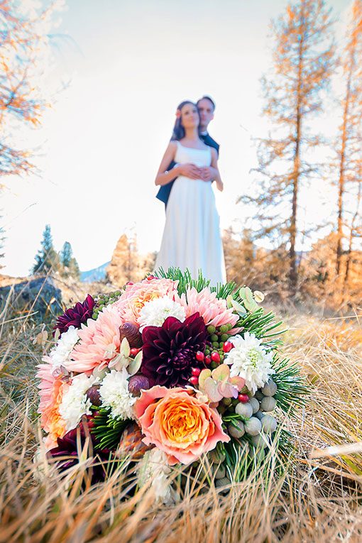 Mountain Wedding. Junges Brautpaar steht auf einer Anhöhe am Berg im Herbst. Der Bräutigam steht hinter der Braut und umarmt sie. Im Vordergrund das Hochzeitsdetail: der Brautstrauß in leuchtend bunten Herbstfarben mit Tannenzweigen.
