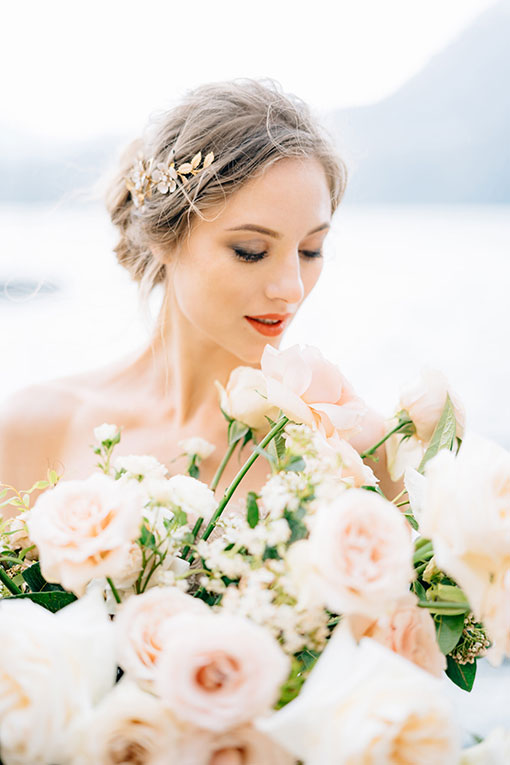 Junge Braut mit schönem Hochzeit Make-up, lockerer Hochsteckfrisur und zartem Haarschmuck aus goldenen Blüten und Blättern steht mit Brautstrauß aus zartrosa Rosen am Ufer eines Sees.