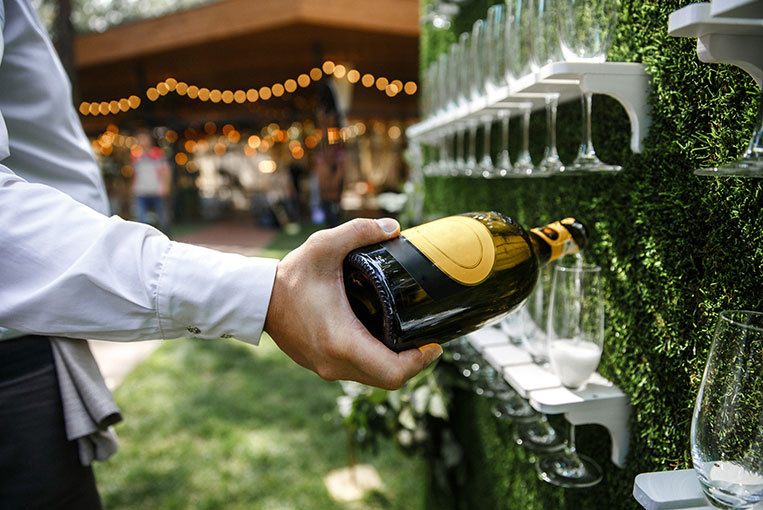 Champagnerwand für ein Event oder Hochzeit. Outdoor Service Caterer steht im Freien und schenkt eine Flasche Champagner in Gläser, die an einer Wand mit Kunstgras festgehalten sind. Im Hintergrund Gäste.