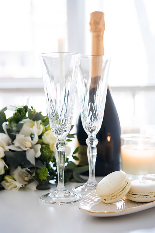 Stilvolles Ambiente mit Champagner, Kristallgläsern und einer Schale mit Maccarons, die auf einem Tisch dekoriert sind. Im Hintergrund Kerzenlicht und Blumen.