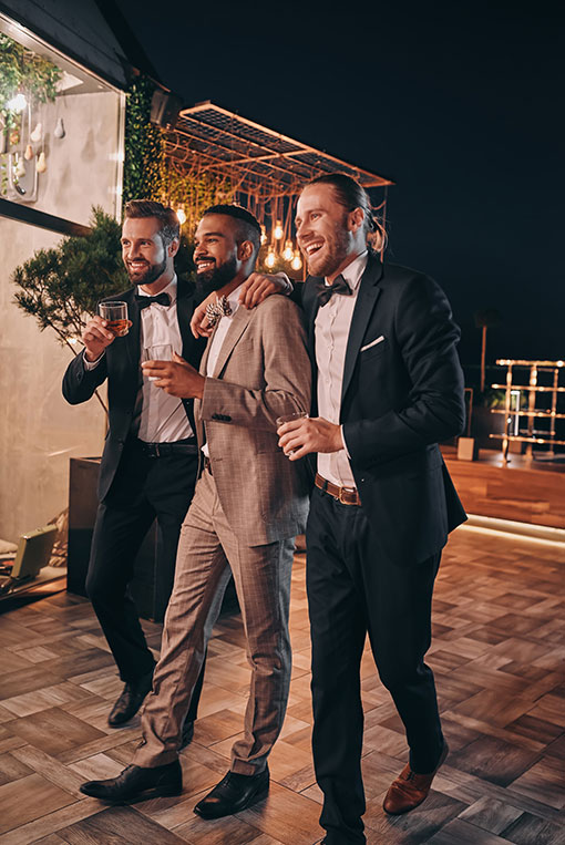 Drei gut aussehende Männer in Anzügen und Fliege trinken Whiskey und unterhalten sich, während sie Zeit auf einer Party verbringen.