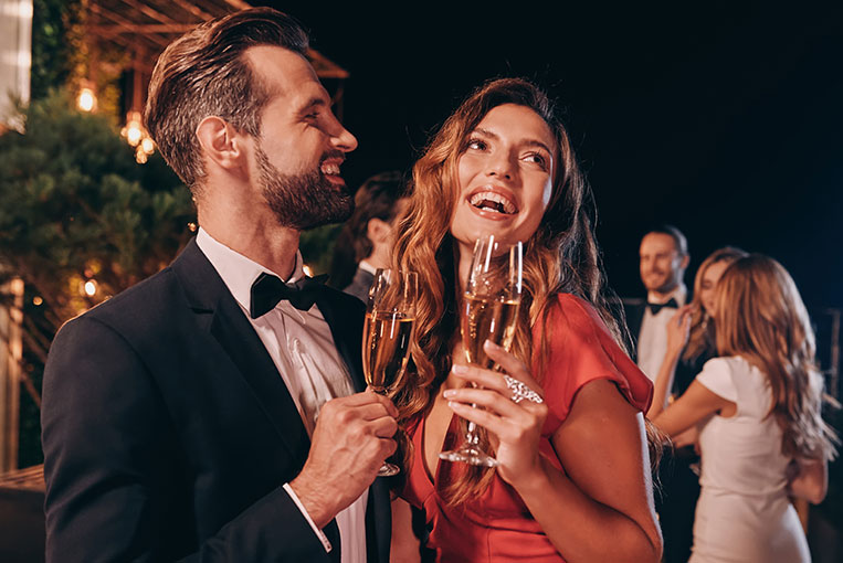 Hübsches junges Paar in eleganter Festkleidung stößt auf einer luxuriösen Sommerparty mit Champagner Gläsern an
