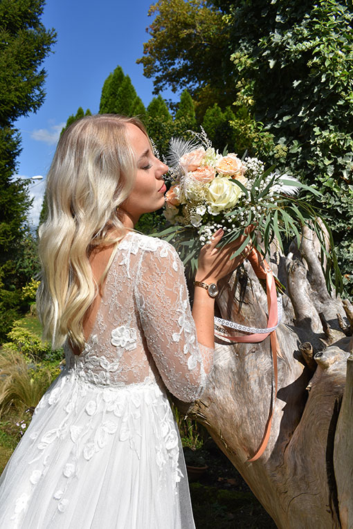 Hochzeitsportrait einer jungen Braut. Sie steht mit geschlossenen Augen an einer Baumwurzel in einem Garten und hält den Brautstrauß mit Federn und langen Bändern in der Hand. Hochzeitsdekoration. Hochzeitsaccessoires.