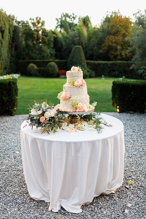 Hochzeitsdekoration. Hochzeitstorte mit Blumendekor steht auf einem runden Tisch mit weißer Tischdecke im Innenhof eines Gartens.