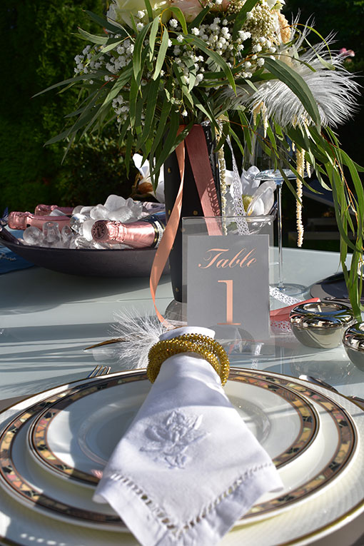 Tischdekoration für eine Hochzeit oder Event. Tischgedeck mit Stoffservietten. Tischnummer in Acrylrahmen. Teelichthalter aus Silber. Blumendekor. Im Hintergrund eine Schale mit Champagner Flaschen auf Eiswürfeln.
