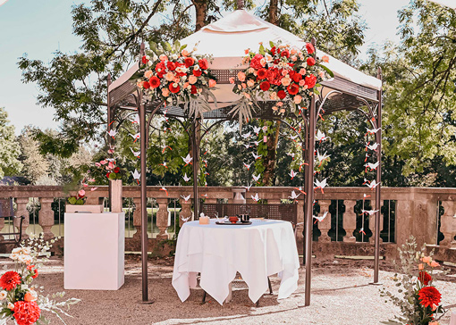 Junge Braut mit schönem Hochzeit Make-up, lockerer Hochsteckfrisur und zartem Haarschmuck aus goldenen Blüten und Blättern steht mit Brautstrauß aus zartrosa Rosen am Ufer eines Sees.